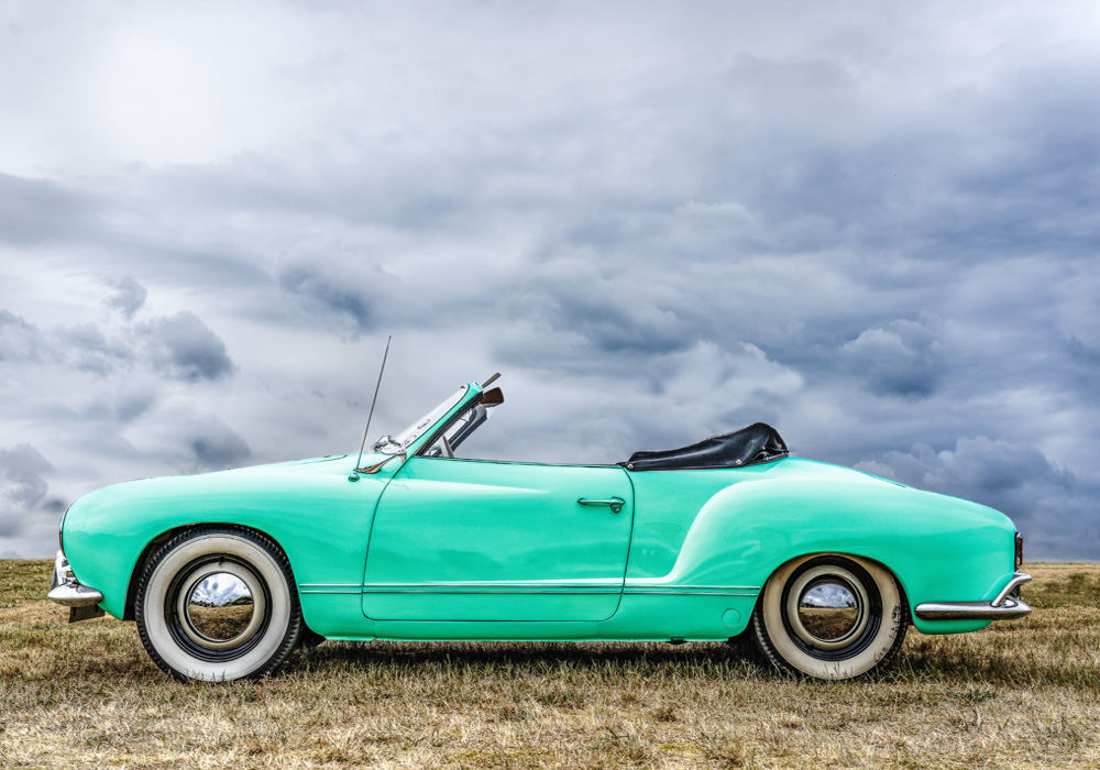 Karmann Ghia convertible in blue parked on a grassy field under dramatic cloudy skies