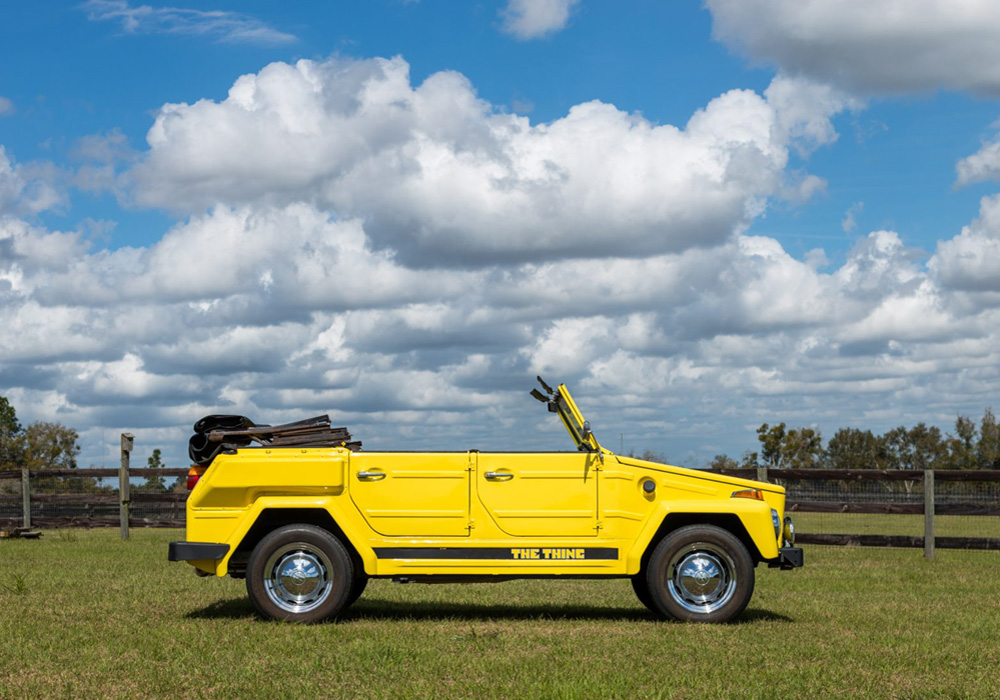 Volkswagen Thing in bright yellow parked on grass with trees and a wooden fence in the background